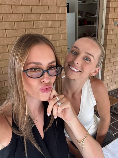 Two women pose closely together in a selfie-style image. The woman in the foreground wears black-rimmed glasses and a black top, with light brown hair. The woman behind her has her blonde hair pulled back and is wearing a white tank top. Both are smiling. A gold bracelet adorns the arm of the blonde woman. Behind them, a brick wall can be seen. In the upper left corner, a glimpse of the inside of a house is visible.