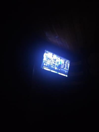 The image shows a dimly lit room with a television screen displaying a brightly lit image. The television is positioned at an angle, making the content difficult to discern clearly, though it appears to be a scene with people and some sort of structure, possibly outdoors with trees. The rest of the room is predominantly dark, with a hint of wooden paneling visible at the top of the frame, suggesting a ceiling or wall. The overall impression is of a domestic setting at night, with the television being the primary source of light. There is no discernible information within the image to indicate the location in the city of Sovetsk, Russia.