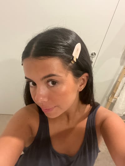 A young adult female is depicted in a close-up, indoor setting within Lordelo do Ouro e Massarelos, Portugal. She has dark hair parted down the middle, brown eyes, and a tanned complexion, looking directly towards the viewer with a subtle smile. A light pink, spade-shaped hair clip with gold-colored metal accents is fastened to the right side of her head, above her ear. A small, circular gold-colored stud earring is visible in her right earlobe. She is wearing a dark-colored tank top. The background consists of a light-colored wall, a segment of a white door with a silver-toned handle to the right, and a multi-tiered wooden rack holding light-colored towels positioned further to the right. The environment appears to be a domestic bathroom or dressing area.