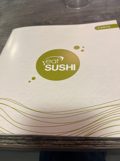 The image shows an "Eat Sushi" restaurant menu placed on a dark-colored table. The menu, oriented vertically from the viewer's perspective and photographed from a slightly overhead angle, features an off-white background. A large, circular lime green logo is centered, containing the white text "Eat SUSHI." The word "Eat" is stylized and positioned above "SUSHI." Small, lime green dots are scattered around the main logo.
In the upper left corner, a vertical lime green tab displays the word "CARTE," which means "menu" in French. The right side of the menu cover is decorated with several thin, wavy lime green lines. Part of a clear drinking glass is visible on the table surface to the left of the menu. The table itself has a dark, textured appearance, possibly wood or laminate. The scene suggests an indoor dining setting, likely a sushi restaurant in Andrézieux-Bouthéon, France, where the menu is presented for ordering. No people or specific activity beyond the presence of the menu are depicted. The time of day and weather are not discernible.