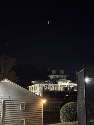 A nighttime scene depicts the exterior of a building complex illuminated by lights. The architecture features white trim and multiple levels, possibly apartments or condos. A fence runs along the upper edge. Visible against the dark sky are a few faint light streaks and a small bright object, suggesting stars or distant lights. A dark bush and the bare branches of a tree provide contrast. The image exhibits a dark and quiet ambiance.