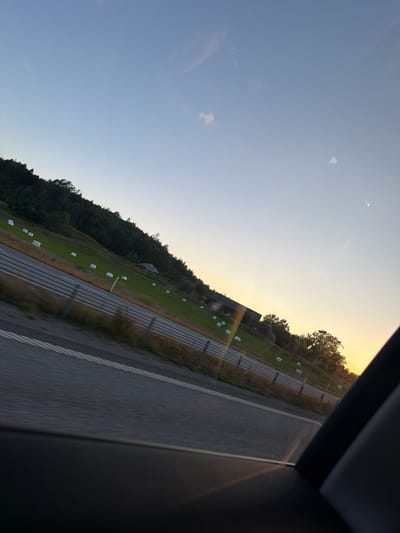 The image, captured from a vehicle, depicts a rural landscape in Munkedal, Sweden, during what appears to be sunset. The sky is a gradient of pale blue to pale yellow near the horizon. A sloped green field, dotted with white wrapped hay bales, stretches across the midground. A dark line of trees borders the top of the field. A road with white lane markings runs diagonally across the foreground, with a fence separating it from the field. In the distance, a building complex with a dark roof is visible on the field. The lower right portion of the image shows the interior of a car, likely a window frame. The overall scene suggests a serene countryside view experienced during travel.