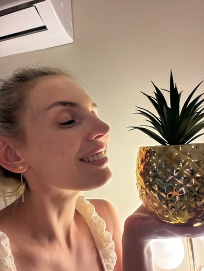 A young woman with blonde hair pulled back in a bun, wearing a white lace top and star-shaped earrings, is holding a gold pineapple-shaped pot with a small plant in it. She is looking down at the pot with a smile. The background is a plain wall with an air conditioning unit in the upper left corner and a light fixture in the lower right. The image is lit with warm, soft lighting. The scene takes place in a residential setting in Mojanovići, Montenegro.