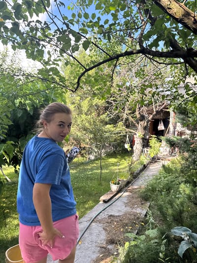 The image depicts a young woman in a backyard setting in Balivka, Ukraine. She is wearing a blue t-shirt and pink shorts. The woman is holding a smartphone in her right hand, and she is looking towards the camera with a surprised expression. Her left hand is resting on her leg.
The scene is an outdoor garden with lush green foliage. A pathway made of concrete runs through the yard, and a garden hose is visible alongside it. In the background, there is a brick structure that appears to be a small shed or outbuilding. Hanging plants are visible near the structure, and a white bucket with an orange flower is placed on the pathway. The time of day appears to be daytime, and the weather seems pleasant and sunny, with dappled sunlight filtering through the trees. The overall impression is one of a casual, outdoor activity in a residential area. No visible text is present in the image.