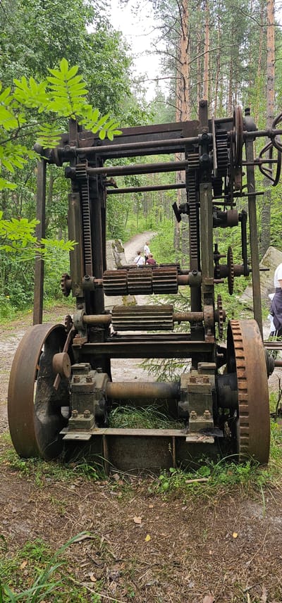 In Medvezhyegorsk, Russia, an abandoned industrial machine, likely from the Soviet era, rests amidst a forest. The machine, characterized by its large gears, rusted metal frame, and weathered components, stands on a dirt path. The surrounding environment is a dense forest with tall pine trees and lush green foliage. In the background, two individuals are visible walking along the path, adding a sense of human presence to the scene.