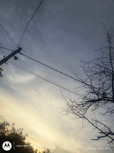 This is a low-angle outdoor photograph of a telephone pole against a cloudy sky. The sky is a mix of dark gray clouds on the left, transitioning to lighter, brighter clouds with a yellowish hue on the right, suggesting it may be late afternoon or early evening. The bare branches of a tree extend into the upper portion of the frame from the left. A telephone pole with several wires attached is visible on the left side of the image. A street light is attached to the pole. The image appears to have been taken in Melnytsia-Podilska, Ukraine.
Visible text overlays indicate the date and time as "30 черв. 2026 р., 20:47" (June 30, 2026, 20:47) and "ЗНЯТО НА motorola edge 40 neo" (SHOT ON motorola edge 40 neo). A Motorola logo is also present in the lower right corner. No people are visible in the image.