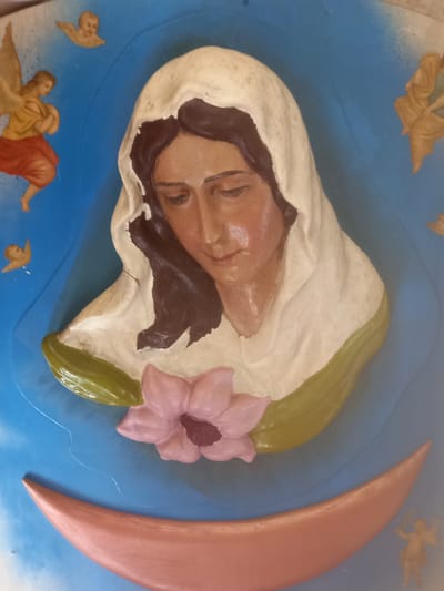 This is a close-up, overhead shot of a religious relief sculpture. The central subject is a serene-looking woman, likely depicting the Virgin Mary, wearing a white veil that covers her head and shoulders. Her eyes are closed, and her face has a peaceful expression. Dark hair is visible around her face, peeking from under the veil. A pink flower with a dark center rests near her head. To the right of the central figure, a large, pale pink crescent moon is visible. The background is a vibrant blue, possibly representing the sky or heavens.
Scattered around the central figure are smaller angelic figures, depicted in a somewhat stylized manner. One angel, with golden wings and an orange garment, is positioned in the lower left corner, appearing to be reaching out. Another angelic figure, also in golden tones, is seen in the upper left corner. A smaller, cherubic figure is located near the bottom center of the image, and another similar figure is in the upper right. These figures suggest a celestial or divine setting. The overall impression is one of peace and spiritual devotion. The sculpture appears to be made of a solid material, possibly plaster or resin, and has been painted in various colors. The scene evokes a sense of timelessness and religious reverence.