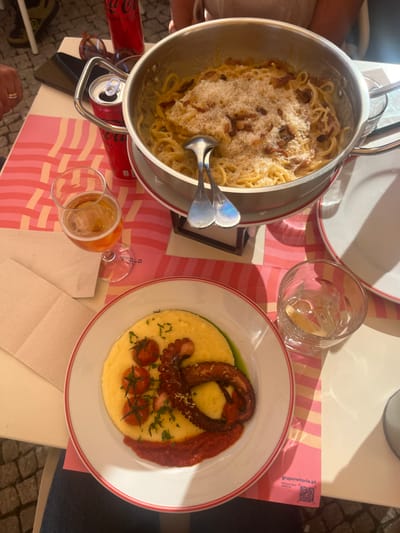 An overhead view captures a dining table in an outdoor setting within Lordelo do Ouro e Massarelos, Porto, Portugal. The table features pink and white striped placemats, one displaying "gruporeitoria.pt" and a QR code. A large stainless steel pot on a stand holds spaghetti carbonara, generously topped with grated cheese and bacon, accompanied by metal serving tongs. A pilsner glass containing amber liquid (likely beer) and two red Coca-Cola cans (one open, one closed) are positioned nearby. Another white plate with a red rim holds a meal consisting of yellow polenta, a cooked octopus tentacle, several halved cherry tomatoes, fresh green herbs, a portion of red sauce, and a drizzle of green oil. A clear glass with water and a lemon wedge is also on the table. A black smartphone and brown sunglasses rest on a placemat. Partially visible are the hands and arms of at least two individuals seated at the table. The ground below the table appears to be cobblestone. Direct sunlight casts distinct shadows across the scene.