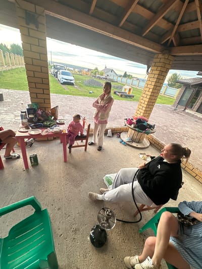 In the city of Peplayaeva, Russia, under a wooden-roofed open-air structure with brick pillars, a group of people are gathered. In the foreground, two individuals are seated on chairs, one of whom is smoking a hookah. To the left, a small pink table is laden with food and drinks, attended by a young child seated on a wooden chair. Further back, a woman stands with her arms crossed, looking towards the left. The ground is paved with concrete. In the background, a green lawn stretches out towards a fence, with houses and trees beyond. Two cars are visible parked on a dirt road. The sky is overcast.