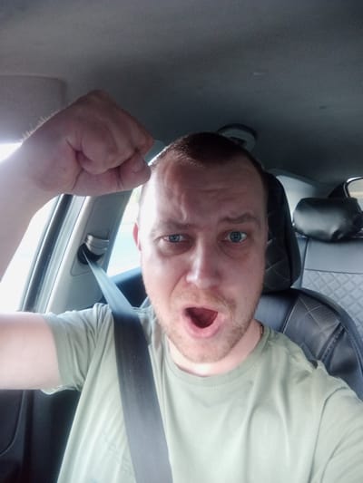This media file shows a man inside a vehicle, likely a car. He is wearing a light green t-shirt and a seatbelt is visible across his chest. His head is tilted, and his mouth is open in an expression that could be surprise or excitement. His eyes are blue and appear to be looking at the camera. A fist is raised in the upper right portion of the frame, suggesting an energetic or emphatic gesture. The interior of the car is visible, including a car seat with a diamond-stitched pattern and a headrest. The scene appears to be indoors, within the confines of the vehicle. The lighting suggests it might be daytime, possibly sunny, given the bright highlights. There is no visible text in the image. The overall impression is a spontaneous selfie taken during a car ride.