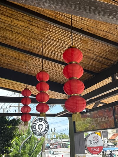The image shows an outdoor covered area with a bamboo ceiling. Red paper lanterns are hung in strings, with one string displaying four lanterns stacked vertically and another showing two lanterns. A sign for "MOTIVE COTTAGE RESORT" is visible, along with signage for "STREET FOOD" and "Coca-Cola" with Thai script. In the background, there is a street scene with buildings, vehicles, traffic lights, and trees, suggesting a commercial area in Ban Bang Niang, Thailand. The overall impression is of a restaurant or cafe exterior.