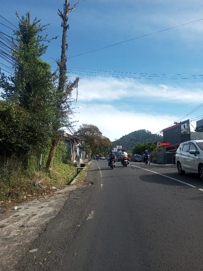 The scene depicts a daytime street view in the city of Phonengam, Laos, under a clear blue sky with sparse white clouds.

On the left side, an unpaved shoulder with scattered debris, including a white plastic cup, borders a concrete drainage ditch. Beyond this, dense green foliage and a tall, bare, moss-covered tree trunk with several utility lines running across the upper frame are visible. Further down the road on the left, a series of simple, single-story buildings with varied facades are partially obscured by vegetation.

The central and right portions of the image feature a two-lane asphalt road. A solid white line delineates the right edge of the traffic lane. Vehicle traffic proceeds away from the viewer; a light-colored SUV is partially visible on the far right. Several motorcycles are present: one rider wears a light-colored top and helmet, another wears a blue shirt and helmet, and additional motorcycles are visible further down the road. A dark sedan is also present in the midground traffic.

On the right side of the road, a dark-colored commercial building with red accents displays a "RedDoorz" sign and a "K1 Hotel" sign. In the background, a large, tree-covered mountain rises prominently.