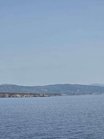 The image displays a calm, blue sea stretching towards a coastline under a clear, pale blue sky. The coastline features rugged cliffs dotted with vegetation and a settlement of buildings. In the distance, rolling hills rise, also sparsely covered with greenery and small structures. A lighthouse with a black and white striped tower is visible on the coast. The water's surface is gently rippled, reflecting the soft daylight. This scene captures the serene coastal landscape of Sol de Mallorca, Spain.