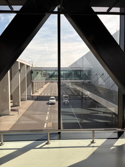 This is a photograph taken from inside a building, likely an airport terminal, looking out through a large window. The view is framed by dark, angular metal beams, creating a triangular opening. Outside, a modern airport scene unfolds. A glass skybridge connects two buildings, casting reflections on the tarmac below. Several vehicles are visible on the road, including a white car and a small utility vehicle. The word "BUS" is marked on the road. The buildings are characterized by concrete pillars and a sleek, modern facade. The sky is partly cloudy. The lighting suggests it is daytime, with distinct shadows cast on the ground. The location is identified as Schönefeld, Germany.