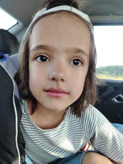 A young girl with dark hair and brown eyes is captured in a close-up shot, looking directly at the camera. She is wearing a white headband and a striped t-shirt. The background suggests she is inside a vehicle, with a window visible on the right side showing a blurred landscape of trees and sky. Sunlight streams in, casting shadows and highlights on her face and clothing. The overall mood is candid and relaxed, possibly during a car journey. The time of day appears to be daytime, given the bright natural light. The location is identified as Tap'iushka, Russia.