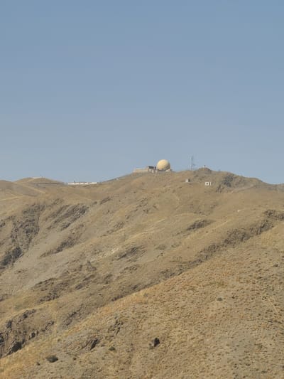 This media file depicts a vast, arid mountain landscape under a clear blue sky, suggesting it's daytime. The camera is oriented vertically, showing a steep, rocky mountainside covered in sparse, dry vegetation. Several structures are visible perched on the mountain's crest or on ledges.

The most prominent object is a large, light-colored dome, likely a radar or weather station, situated on the upper part of the mountain. Beside it, there are smaller rectangular buildings and a tall lattice tower, possibly a communication antenna. Further along the ridge, a long, white building complex is visible. These structures suggest a research, communication, or monitoring facility located in a remote, elevated area.

The scene is barren and rugged, characterized by sharp slopes, rocks, and the dry, yellowish-brown terrain typical of a desert or semi-arid mountain region. The sparse vegetation hints at a dry climate. The clear sky indicates fair weather. The location appears to be Zhob, Pakistan, a region known for its mountainous terrain and arid climate. There are no people or animals visible, and no text is present in the image. The overall impression is one of isolation and a functional, possibly scientific, outpost in a harsh natural environment.