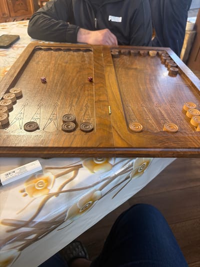 A backgammon board is positioned on a table, with a person seated on the left side wearing a black Puma jacket. Another person is seated on the right, wearing denim jeans. Two dice are visible on the lower half of the backgammon board. The table is covered with a white tablecloth adorned with a floral design. A barcode sticker or label is also present on the table near the board. The image provides no verifiable data for the location Dimitrov, Armenia.