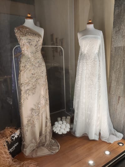 This image depicts a shop window display in Thessaloniki, Greece, featuring two mannequins adorned with formal gowns. The mannequin on the left is dressed in a champagne-colored, one-shoulder gown embellished with intricate beadwork and sequins in floral and vine patterns. The gown has a flowing skirt. The mannequin on the right wears a white, sleeveless gown with a similar all-over beading and sequin detail, featuring a sheer overlay that drapes like a cape. In the foreground, to the left, is a black shopping bag with the white text "άρογλου | υφάσματα" (meaning "Aroglou | Fabrics" in Greek) and is surrounded by light pink feathers. To the right of the gowns, a small arrangement of white candles and white floral decorations in a glass bowl is visible. The display rests on a wooden surface, illuminated by spotlights embedded in the surface. The background is dimly lit and features reflections that obscure specific details of the store's interior. The overall impression is one of elegance and formal wear.