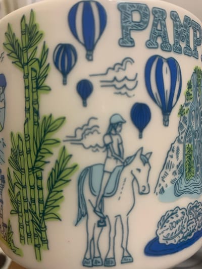 The image is a close-up of a white ceramic mug featuring a whimsical, hand-drawn illustration. The artwork depicts a collection of diverse elements, including several hot air balloons in varying sizes, with bold blue and white stripes. There are also stylized bamboo stalks with green leaves on the left side of the mug. In the center, a young person wearing a cap is riding a horse. To the right, there is a depiction of a rocky landscape with a waterfall or flowing water, and a plate of food, possibly a salad or vegetables, on a blue base. The word "PAMP" is prominently displayed in large, textured, light blue letters at the top of the mug. The overall style is simplistic and illustrative, with a focus on line art and solid color fills. There is no discernable location in Angeles City, Philippines, depicted in this image; the imagery appears to be a generic, decorative theme.