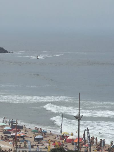 The image depicts a crowded beach scene in Espinho, Portugal, under a hazy sky. The ocean is rough with significant waves crashing near the shore. On the beach, numerous people are sunbathing, relaxing under umbrellas, and walking along the sand. Several surfboards are visible, indicating surfing activity. In the distance, a person is riding a jet ski. The foreground shows a variety of beach umbrellas, chairs, and some flags, suggesting a recreational event or busy beach day. The overall atmosphere is one of leisure and outdoor activity.