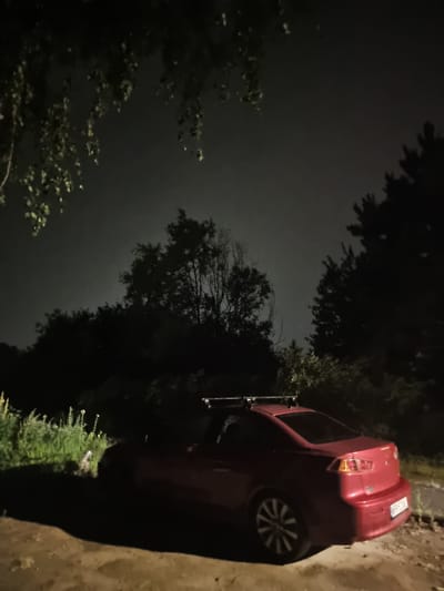 The image depicts a red four-door sedan parked at night on a dirt road or unpaved area. The car has a roof rack installed. The environment is characterized by dark foliage and trees surrounding the scene, with the sky appearing hazy or overcast. The lighting suggests it is nighttime. The location is identified as Szczaki, Poland. There are no people visible in the image.