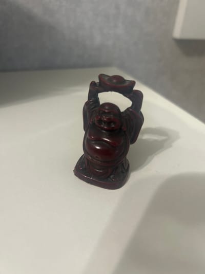 A small, dark red figurine of a laughing Buddha is centered on a white surface. The figurine is depicted with a full belly, a smiling face, and arms raised, holding a round object above its head. The surface appears to be a tabletop or shelf, and in the background, a wall with a textured, gray finish is visible. The lighting casts a soft shadow to the right of the figurine. The location in the city of Kokhma, Russia, is not discernible from the visual information provided.