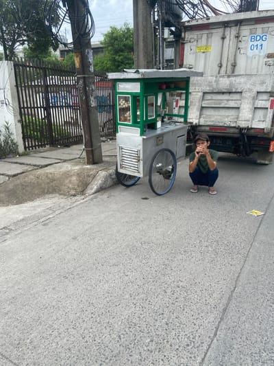 The image shows a street vendor in Bojongsoang, Indonesia (latitude: -6.967, longitude: 107.638). The vendor is sitting on the ground in front of his food cart. He is wearing a blue shirt and a green shirt over it. His back is turned to the camera. He is holding his chin with his left hand. The cart is a silver, two-wheeled cart with a green and white front panel. It has a glass display case on the front. There is a white truck parked behind the vendor and cart. The truck has "BDG U11" written on its side. The street is paved in grey concrete. There is a black power pole to the left of the vendor and a black iron gate with a few small metal panels behind the vendor. The ground is covered in gray pebbles. There are no other people in the scene. The sky is partially visible in the background. The image was taken during the day in what appears to be dry conditions.