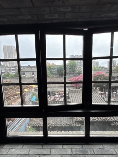 This image captures an exterior view from a gridded, dark-framed window, situated within an interior space characterized by a grey brick wall above and below the window. The scene depicts a mixed urban environment in Qingyang District, China, transitioning from traditional architecture to modern high-rises under an overcast sky.

Through the window panes, the midground reveals a complex of traditional Chinese-style buildings featuring dark grey tiled roofs with curved eaves. Several such structures are visible, including one prominent grey-brick building with multiple windows and two visible air conditioning units mounted on its facade. To the left, a wooden trellis-like structure extends over an outdoor area. Numerous people are present in a pedestrian area below and between the traditional buildings, forming several clusters and appearing to walk or stand. Many individuals are dressed in light-colored clothing. A vibrant tree with abundant pink or fuchsia flowers stands out amidst green foliage, positioned centrally within the traditional complex. A blue sign with white script, partially obscured, is also visible.

In the background, beyond the traditional structures and trees, several tall, modern high-rise buildings rise, characterized by numerous windows and distinct architectural styles, offering a stark contrast to the foreground. The sky is uniformly grey and overcast. The window glass itself exhibits minor smudges or dust.