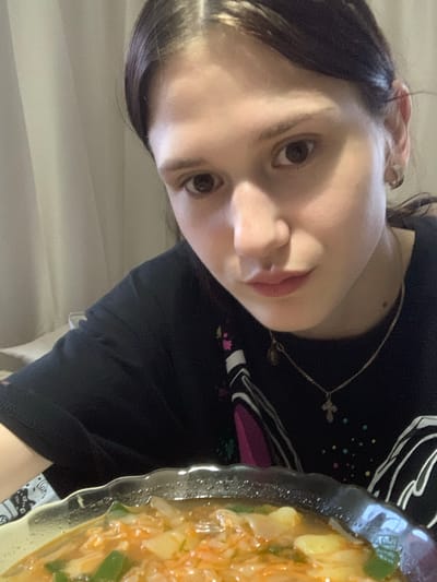 A young woman with dark hair pulled back is positioned in the foreground, looking towards the camera. She is wearing a black t-shirt with a colorful abstract pattern. A silver cross necklace is visible on her neck. To her right, a clear bowl filled with a vibrant orange soup containing vegetables and noodles is visible. The background consists of a pale, textured curtain. The scene appears to be indoors, possibly a domestic setting. The image was captured on February 3, 2024.