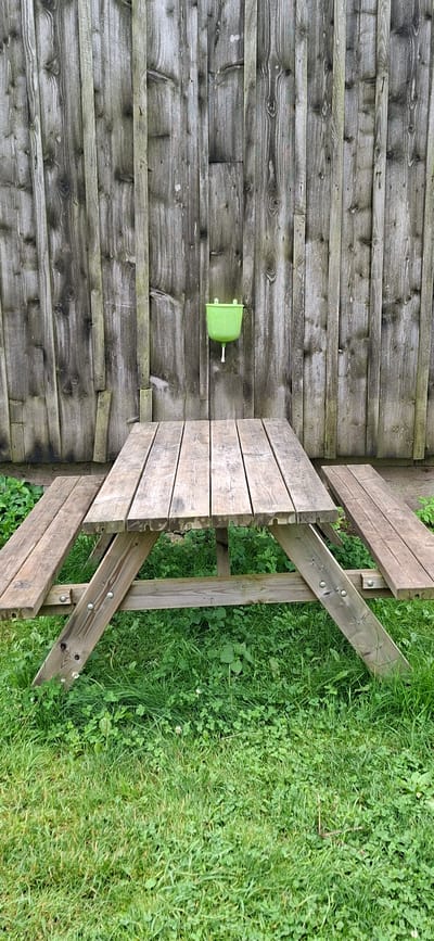 The image shows a rustic wooden picnic table with attached benches set on a grassy area in front of a weathered wooden fence. A small, bright green plastic basin with a white spout is mounted on the fence above and behind the table. The fence is made of vertical wooden planks of varying widths and tones, exhibiting signs of age and weathering. The grass is lush and green, with some clover interspersed. The overall scene suggests an outdoor, possibly rural or park-like setting. The location is stated to be in the city of Kata, Estonia. There are no people or animals visible in the image.