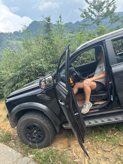 A man is seated in the driver's seat of a black SUV with an open door, parked in a natural environment with dense vegetation. The SUV appears to be slightly tilted on uneven ground. The surrounding area is a hilly terrain with green foliage, possibly indicating a mountainous region. The mirror reflection shows two people standing outside the car. The image was taken near Suối Giàng, Vietnam.