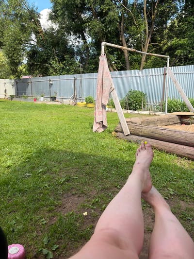 The image captures a person's legs and feet resting on a grassy area, likely outdoors in a backyard or garden setting. The scene is set against a backdrop of trees, a corrugated metal fence, and wooden logs. A patterned umbrella lies partially unfurled on the grass. The sunlight suggests it is daytime, and the lush green grass indicates pleasant weather. The focus on the person's lower extremities, with painted toenails, implies a relaxed, casual moment. The presence of trees and a fence suggests a semi-rural or suburban location. The provided location context of Kimry, Russia, further grounds the scene in a specific geographical area. There is no visible text in the image.