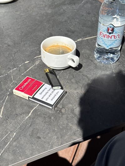 The image depicts a dark marble-patterned table with several objects on it, suggesting an outdoor setting in Jermuk, Armenia. In the center of the frame, a white ceramic coffee cup contains a dark liquid, likely coffee. To the right of the cup, a clear plastic bottle of water is positioned. In the foreground, to the left of the cup, is a pack of "CHAPMAN" cigarettes, red and white in color, with a superimposed health warning in English. A black disposable lighter rests on top of the cigarette pack. The table surface is illuminated by sunlight, casting shadows from the objects. The background is out of focus, but appears to be a natural or outdoor environment. The overall impression is a casual, possibly recreational moment, with the presence of cigarettes and alcohol suggesting adult activity.