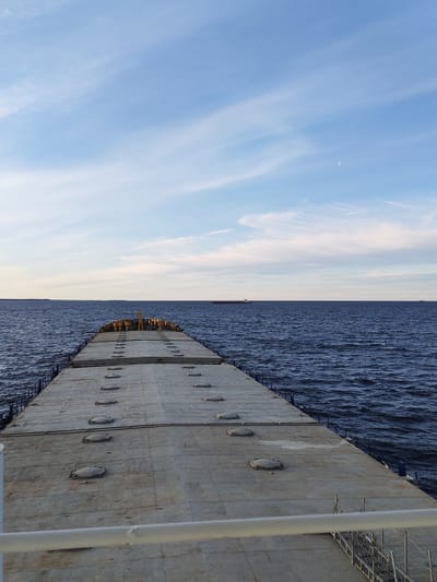 View from a barge or large boat with a concrete deck, showing a body of water and horizon under a blue sky with wispy clouds and a crescent moon. The deck of the boat has several circular manhole covers. There are other ships in the distance. The image appears to be taken at or near dusk. This scene is not in Novaya Svobodka, Russia. There are no people visible in the image.