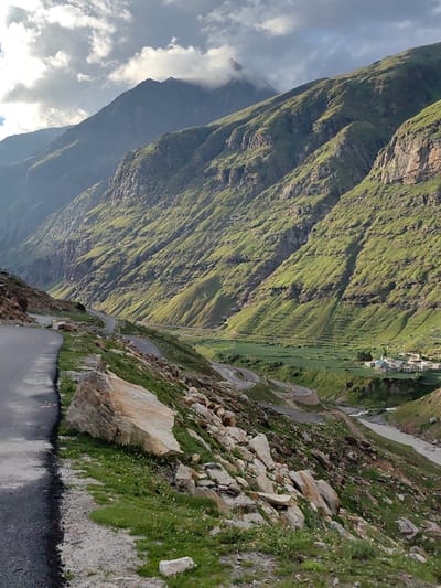 The image captures a sweeping vista of a mountainous region, likely the city of Yari-Khoksar, India, showcasing a dramatic landscape with a winding road and a distant village nestled in a valley. The foreground features a paved road curving to the left, bordered by a steep, rocky embankment covered in patches of green grass and scattered white wildflowers. Large, light-colored boulders are prominent along this embankment.

The mid-ground reveals the road continuing its serpentine path down into a lush green valley. This valley is characterized by terraced fields, suggesting agricultural activity. A river, appearing somewhat murky, flows through the valley floor, with a small cluster of buildings, a village, visible on the far bank. The architecture of the village appears to be traditional, with multi-story structures.

The background is dominated by immense, green-covered mountains that rise steeply from the valley. The slopes are rugged and etched with natural contours and some man-made terraces. The upper reaches of the mountains are partially obscured by dramatic, heavy clouds, with a peak visible piercing through the cloud cover. The lighting suggests it's either early morning or late afternoon, with the sun casting shadows and highlighting the textures of the mountainside. The overall atmosphere is one of rugged natural beauty and a remote, secluded settlement.