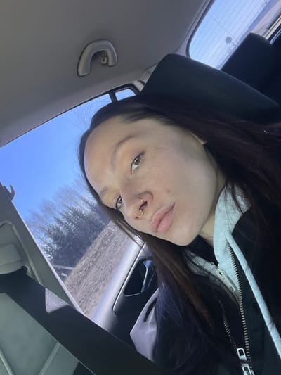 A person is seated in a car, with their head tilted towards the camera. The car interior is visible, including the headrest, rearview mirror, and seatbelt. Outside the window, trees and a blue sky are visible. The person has dark hair, light skin, and a nose piercing. No information indicates that the image was taken in Иж-Бобья, Russia.