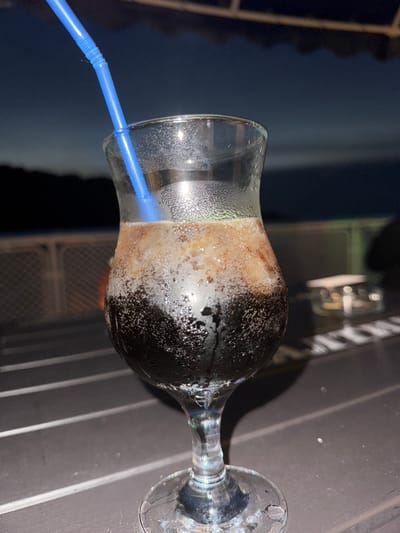 A close-up, low-angle shot features a dark beverage in a stemmed glass with a blue straw. The glass is sweating, and the drink has a foamy head. The beverage appears to be a soda or a similar carbonated drink, possibly a cola, with ice or a frozen component. The setting is outdoors at dusk, suggested by the dark sky and the dimly lit background. In the background, there is a blurred fence and what appears to be a body of water. The table the glass is on is dark and appears to be made of wood or a similar material with visible lines. There are no people clearly visible in the image. The scene suggests a relaxed evening outdoors, possibly at a cafe or restaurant by the water. The location is specified as Makarovo, Russia.