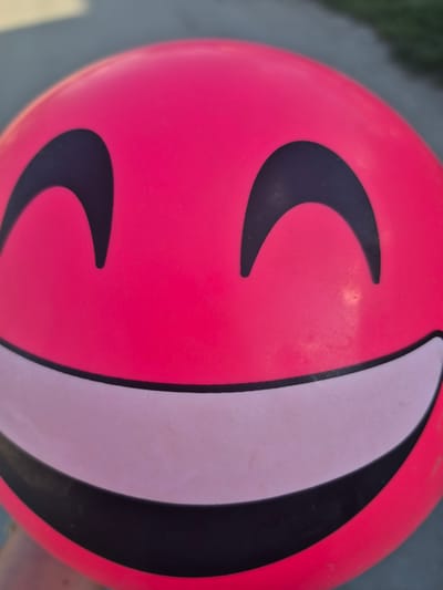 A close-up shot focuses on a pink ball featuring a graphic design suggestive of a smiling face. The "eyes" are depicted as dark, curved shapes, and a large, sweeping curve on the right side, bordered in black and filled with a lighter pink or white hue, represents the "mouth" or cheek. The background is slightly out of focus and appears to show concrete or pavement, possibly indicating the ball is outdoors, potentially in a public space. There are no people visible in the frame. The specific location of Slivo Pole, Bulgaria cannot be determined from the image.