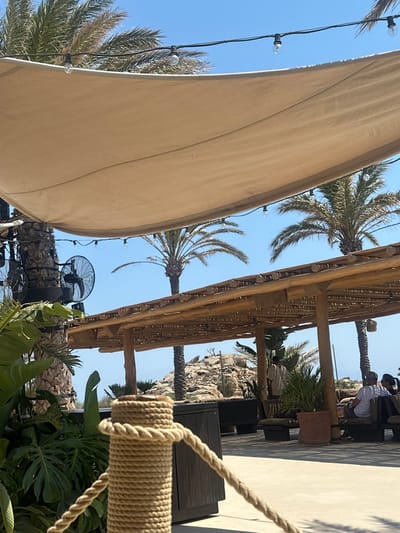 The image depicts an outdoor leisure area in Psarou, Greece, under a clear blue sky. In the foreground, a prominent post wrapped in thick natural fiber rope serves as part of a barrier, with a matching rope extending horizontally. Lush green tropical foliage, including large-leafed plants, borders the immediate left.
The midground features a large, light-beige fabric canopy suspended overhead, with string lights strung beneath it. To the left, an industrial-style black fan is mounted on the trunk of a palm tree. A substantial wooden pergola structure, constructed from natural logs with a slatted roof, occupies the central and right midground. Various potted plants are interspersed throughout this area.
Within the pergola, three individuals are present. One person, wearing a light-colored tunic, stands centrally. To the right, two individuals are seated on wooden benches or low furniture, facing away from the camera; one appears to be a male in a light shirt, and the other a female with dark hair.
The background reveals several tall palm trees, a large rocky hill or formation, and a faint horizon line suggesting the presence of the sea on the far right. The ground throughout the visible area appears to be paved. The scene suggests a sunny day in a relaxed, shaded outdoor setting.