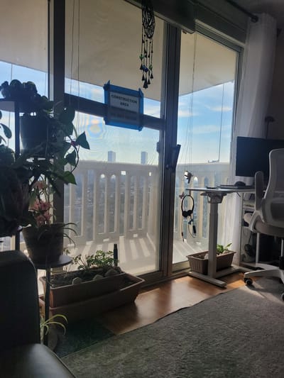 In Fort Lee, United States of America, a sunlit apartment living room features a large window overlooking a city skyline. Through the window, a balcony is visible, cordoned off with a blue sign reading "CONSTRUCTION AREA BALCONY CLOSED DO NOT ENTER." Inside, a desk with a computer monitor and an office chair is set up near the window, suggesting a home office. Several potted plants are placed around the room, adding a touch of greenery. The overall scene conveys a sense of domesticity with a functional workspace, accented by the city view and the unusual detail of the construction warning.