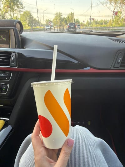 The image shows the interior of a vehicle. A hand holds a white disposable cup with orange and red branding, containing a beverage and a straw. The dashboard is visible, with ventilation grilles, controls, and an integrated screen or navigation system. Through the windshield, a road can be seen with cars and street lights. Trees are visible along the roadside. Based on the GPS location on the navigation system, the vehicle appears to be located near Сухарево, Russia.