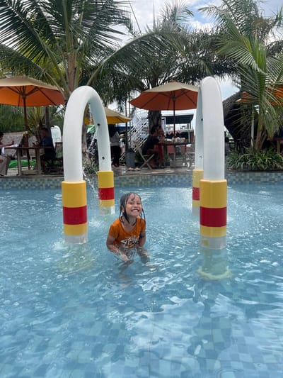 A young female, appearing approximately 5-7 years of age, with dark wet hair, is depicted chest-deep in a light blue tiled pool, smiling broadly. She is wearing an orange t-shirt. Water is spraying from three white, red, and yellow colored arch-shaped water features positioned in the pool behind and to her sides. The background reveals a recreational area with several mature palm trees under a partially cloudy sky. Multiple orange and yellow patio umbrellas provide shade over various groups of individuals seated at tables, suggesting a dining or lounging area adjacent to the pool. Structures include a white staircase leading to an upper level on the right, and various small huts or covered seating areas. The scene is consistent with a water park or resort pool area located in Juwana, Indonesia.