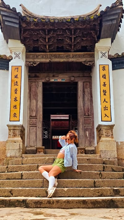 A young woman is sitting on stone steps in front of a traditional building in Sà Phìn Commune, Vietnam. The building has an elaborate wooden facade with Chinese characters on the pillars. The woman is wearing sunglasses, a light blue jacket, green shorts, white socks, and sneakers. She is looking up and holding her hand up to shield her eyes from the sun. The stone steps lead up to the entrance of the building.