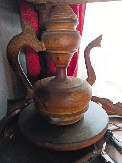This image depicts an indoor scene in Bien Hoa, Vietnam, centered on a carved wooden decorative object resting on a dark, circular wooden base. The object, constructed from dark brown wood with visible grain, exhibits a baluster-shaped body crowned with a finial-like component. It incorporates two carved wooden extensions: an intricate, stylized handle on the left side and a simpler, curved spout-like handle on the right. A layer of fine dust is observable on the object's surfaces and its base. The background contains a section of red, textured fabric and a prominent bright white area on the right, consistent with illumination from a window or strong artificial light. In the lower right foreground, a silver multi-tool with its pliers open and a cluster of white electrical wires secured by a black tie are visible on the supporting surface. No individuals are present, and no actions or interactions are depicted.