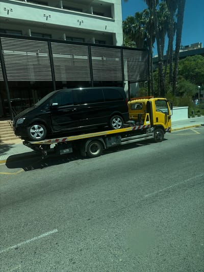 A black Mercedes-Benz Vito van is being transported on the back of a yellow tow truck on a city street in La Pineda, Spain. The tow truck is positioned on the right side of the road, and the van is loaded onto its flatbed. The van appears to be in good condition. The street is paved with asphalt and has yellow lines indicating parking spaces. In the background, there is a modern building with balconies and palm trees. The sky is clear and blue. The overall scene suggests a normal urban environment on a sunny day.