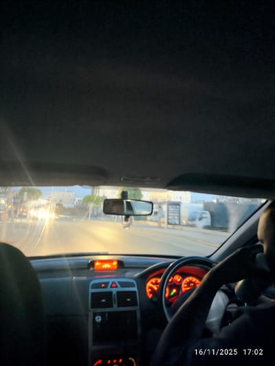 The image depicts the interior of a vehicle, viewed from the front passenger seat looking forward, within the city of Gonyeli, Northern Cyprus / Cyprus. The scene is illuminated by the ambient light of late afternoon or early evening, augmented by vehicle and street lighting.

In the immediate foreground, the dark gray headliner of the vehicle occupies the upper portion of the frame. The dashboard is prominently visible, featuring a digital display centrally located showing "18:24" in orange, and other controls illuminated in red, including air vents and hazard light button. The driver's side of the vehicle is on the right, where the back of a person's head and right shoulder are visible, indicating a driver. The driver's right hand is holding the illuminated steering wheel, consistent with right-hand drive operation. The instrument cluster behind the steering wheel shows red-orange backlighting. A rearview mirror hangs centrally from the windshield, with a small object suspended from it.

Beyond the windshield, a paved road stretches forward, with bright light emanating from oncoming vehicle headlights, causing visible lens flares. Buildings line both sides of the street in the middle distance, interspersed with trees. On the right side of the road, further down, a white truck is visible, and what appears to be a bus stop or roadside structure. The sky above the distant buildings exhibits fading daylight. A timestamp "16/11/2025 17:02" is present in the lower right corner of the image.