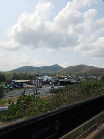 The image depicts a view from what appears to be a moving vehicle, potentially a train. The scene shows a mountain range rising behind a small village or town. The sky is partially cloudy. The town has buildings of varying sizes, a road with power lines, and some vehicles. There are visible utility poles and power lines running along the roadside. The foreground has blurred elements from the vehicle's window or frame. The scene is consistent with a landscape setting in Bang Phra Subdistrict, Thailand.