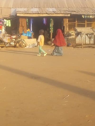 This image captures an outdoor scene in Nunku, Nigeria, likely a village street or market area during either early morning or late afternoon, indicated by the long shadows cast across the dusty, unpaved ground. The light is warm, suggesting clear, sunny weather.
In the foreground, two individuals are walking from left to right. On the left, a young boy, seen from behind and slightly to the side, wears a light yellow long-sleeved shirt, light green trousers, and white shoes. To his right and slightly behind him, another person, whose back is to the viewer, wears a vibrant red head covering, possibly a hijab or shawl, paired with a flowing, patterned dress in shades of blue and white.
The background features a simple, open-front structure with a corrugated metal roof supported by wooden beams. This appears to be a shop or market stall, with various fabrics or clothing items hanging for sale inside. On the far left, a motorcycle or scooter is parked, partially visible. Inside the shop, another figure is dimly discernible. To the right of the main shop opening, there are wooden display stands or shelves, further reinforcing the commercial nature of the building. The overall scene depicts a typical daily moment in a local setting, characterized by basic infrastructure and everyday activity. No text is visible in the image.
