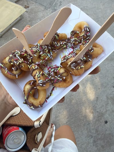 The image shows a close-up view of a white cardboard container holding several mini donuts drizzled with chocolate and sprinkled with colorful jimmies. Wooden skewers are inserted into some of the donuts, suggesting they are being eaten as a snack or dessert. The donuts are golden brown and appear to be freshly fried.
The container is being held by a person whose hands and arms are partially visible. The person is wearing a white top. To the right of the container, a portion of a brown patterned handbag and a red and silver can of Coca-Cola are visible. The background consists of a textured grey surface, likely concrete, with some dappled sunlight and shadows, indicating an outdoor setting. The lighting suggests it is daytime.
The overall impression is of a casual outdoor snack or treat. The context provided indicates the location is Trachoni, Cyprus, hinting at a Mediterranean climate, possibly warm weather. There are no strong emotional cues evident, but the presence of a sweet treat and a beverage suggests a moment of enjoyment or relaxation. The only visible text is on the Coca-Cola can: "330 ml".