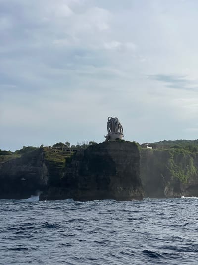 A statue with multiple tubular extensions juts out from a rocky cliff face. Vegetation covers the top of the cliff. Beneath the statue is a structure with vertical supports. A waterfall flows from the base of the cliff into the ocean. The sky is overcast.